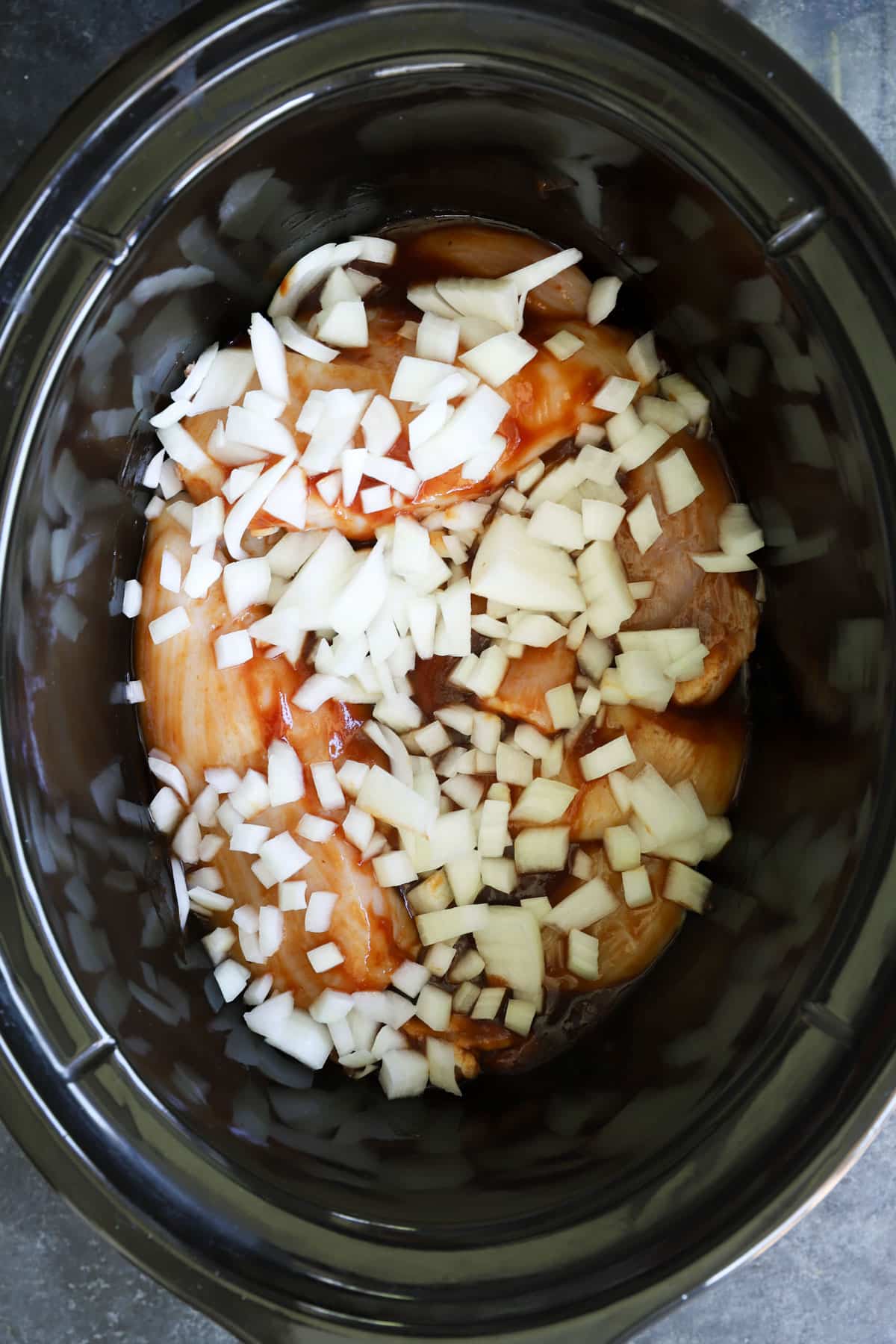 Raw chicken breasts and thighs coated in barbecue sauce topped with chopped yellow onion in a black slow cooker before cooking