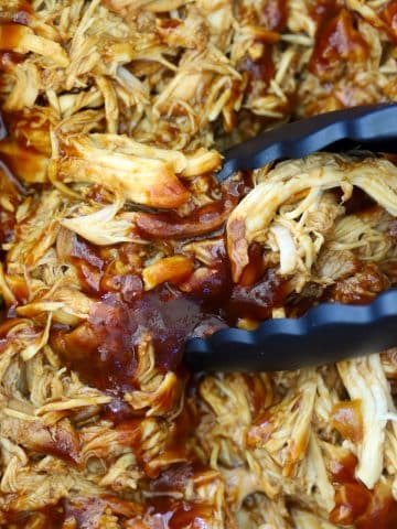 Black tongs stirring saucy shredded barbecue chicken coated in glossy BBQ sauce in a crock pot