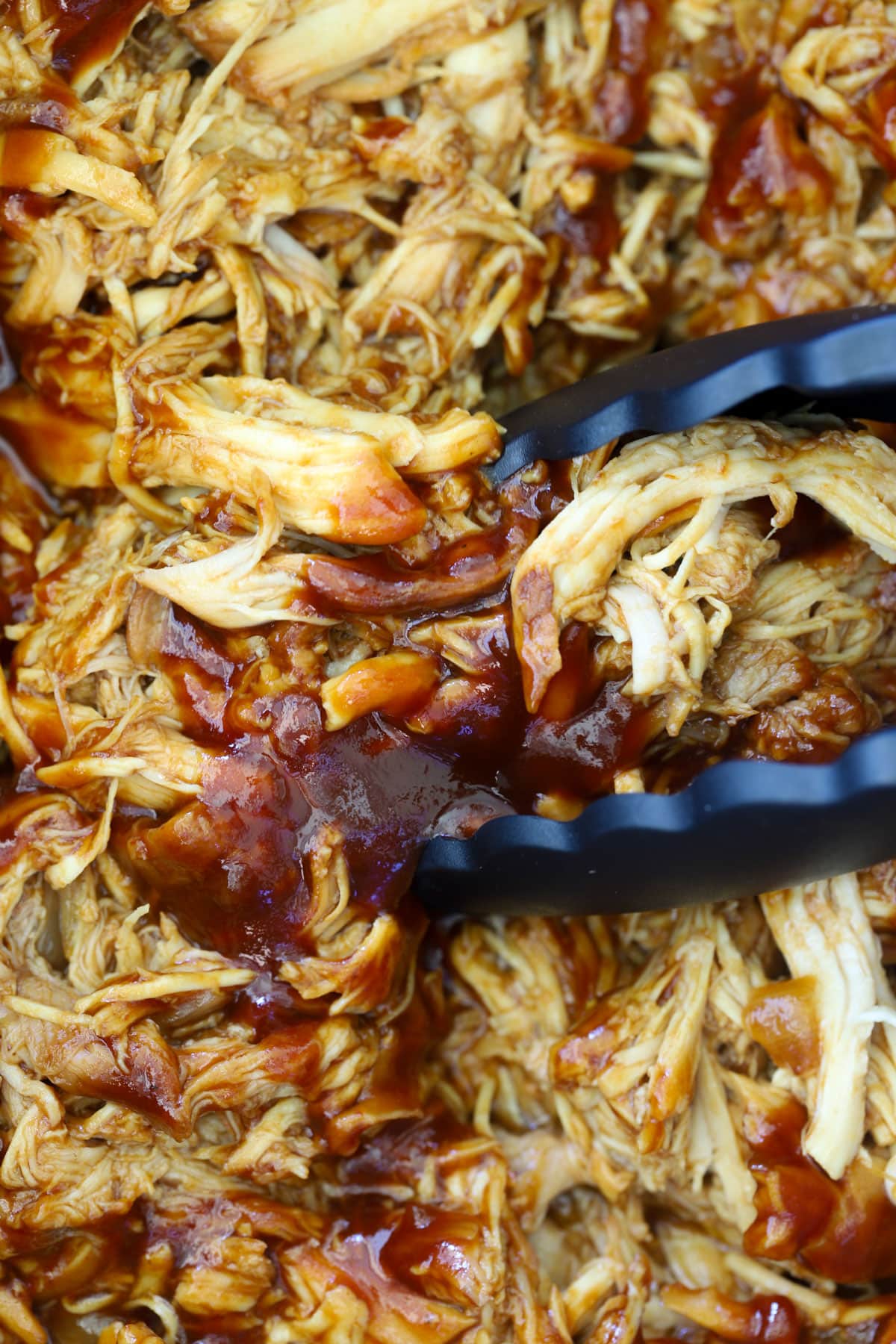 Black tongs stirring saucy shredded barbecue chicken coated in glossy BBQ sauce in a crock pot