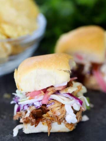 Slow cooker BBQ chicken slider on a soft roll topped with creamy coleslaw and tomato, with a bowl of chips in the background