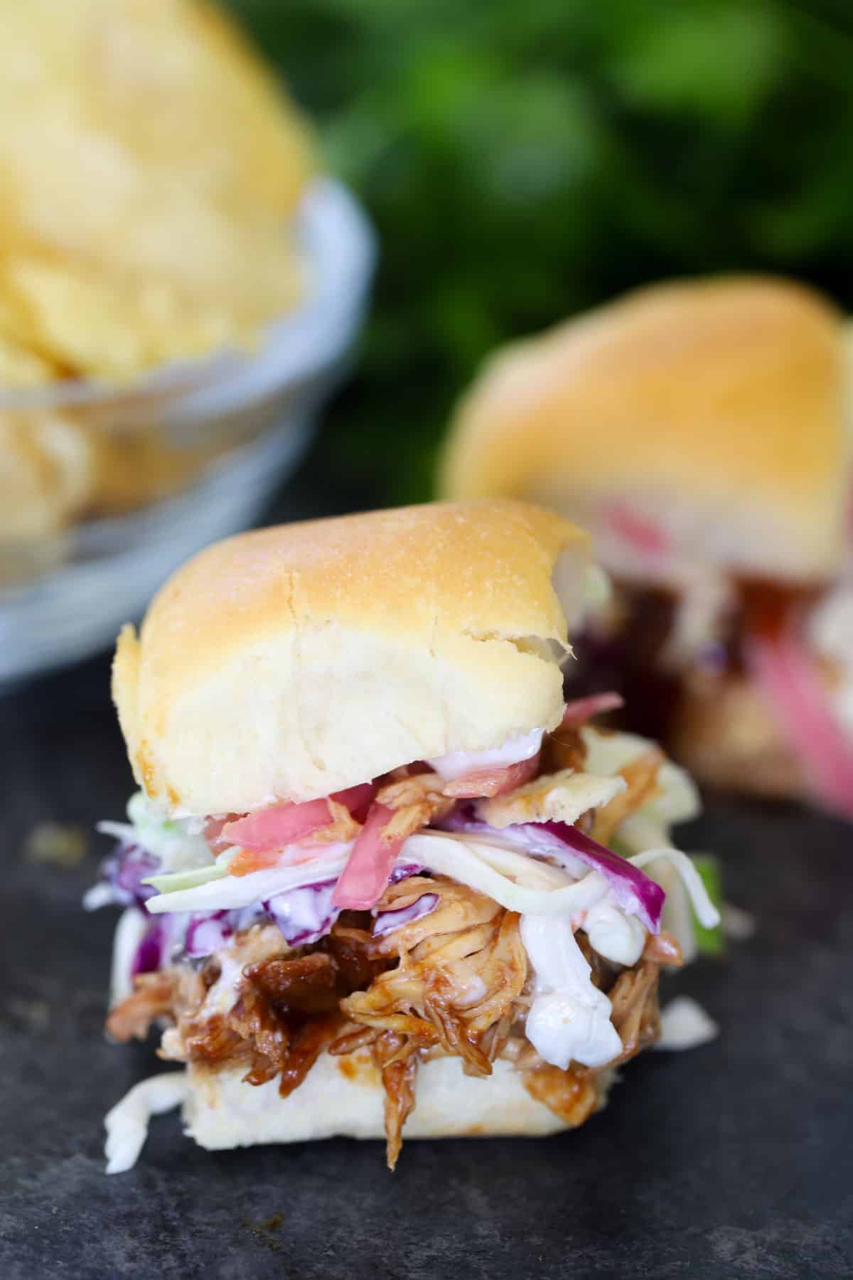 Slow cooker BBQ chicken slider on a soft roll topped with creamy coleslaw and tomato, with a bowl of chips in the background
