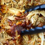 Black tongs stirring saucy shredded barbecue chicken coated in glossy BBQ sauce in a crock pot